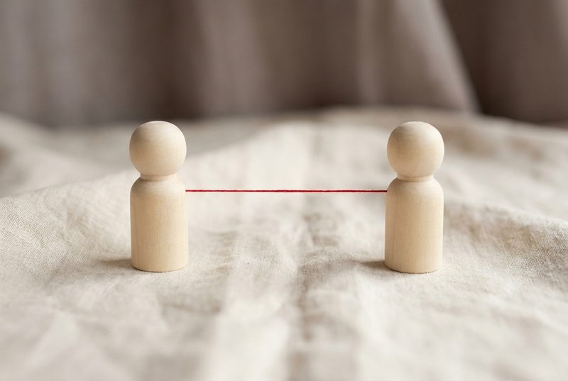 Two plain wooden peg figures standing on linen, facing each other, joined by a single taut red thread.