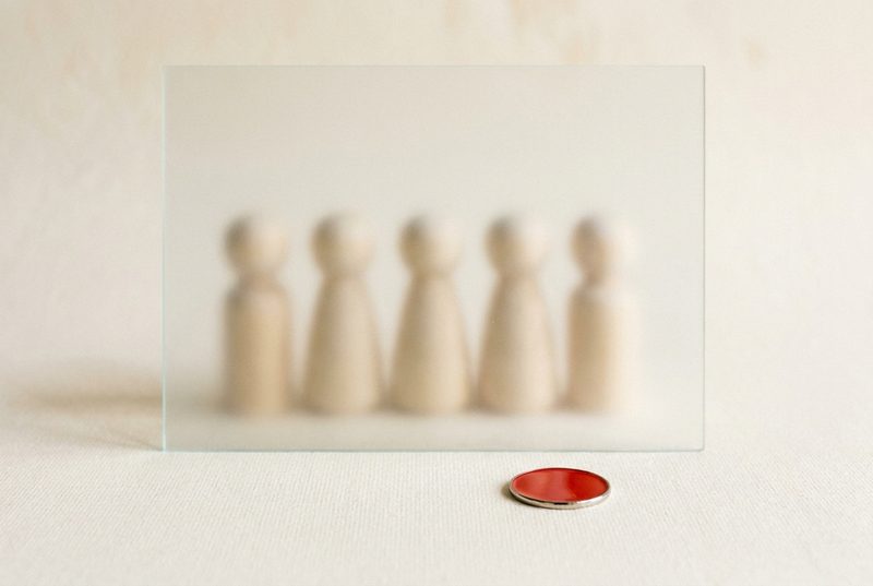 Five wooden figures blurred behind a pane of frosted glass, with a red token lying in front of the glass.